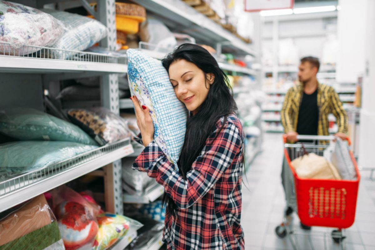 Mulher diante de grande prateleira em interior de loja abraçando um travesseiro, fazendo uso de estratégia de Marketing sensorial que explora o tato ou toque das pessoas no produto