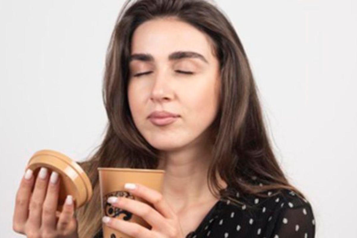 Mulher segura copo de bebida análoga à café e abre sua tampa para sentir cheiro do mesmo, com ar de satisfação e olhos fechados aproveitando estratégia de Marketing sensorial