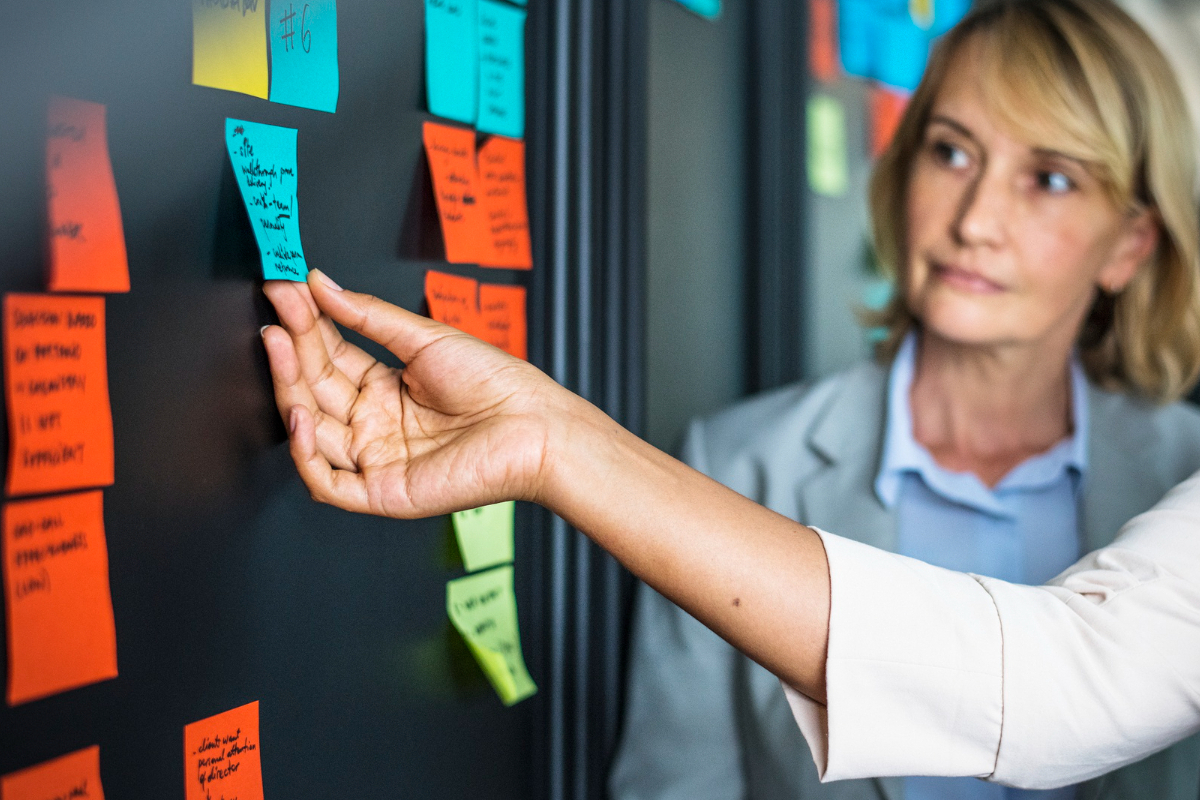 Empresa: Matriz RACI Duas mulheres em empresa analisando anotações em post-its de diversas cores colados em mural escuro para criar Matriz RACI