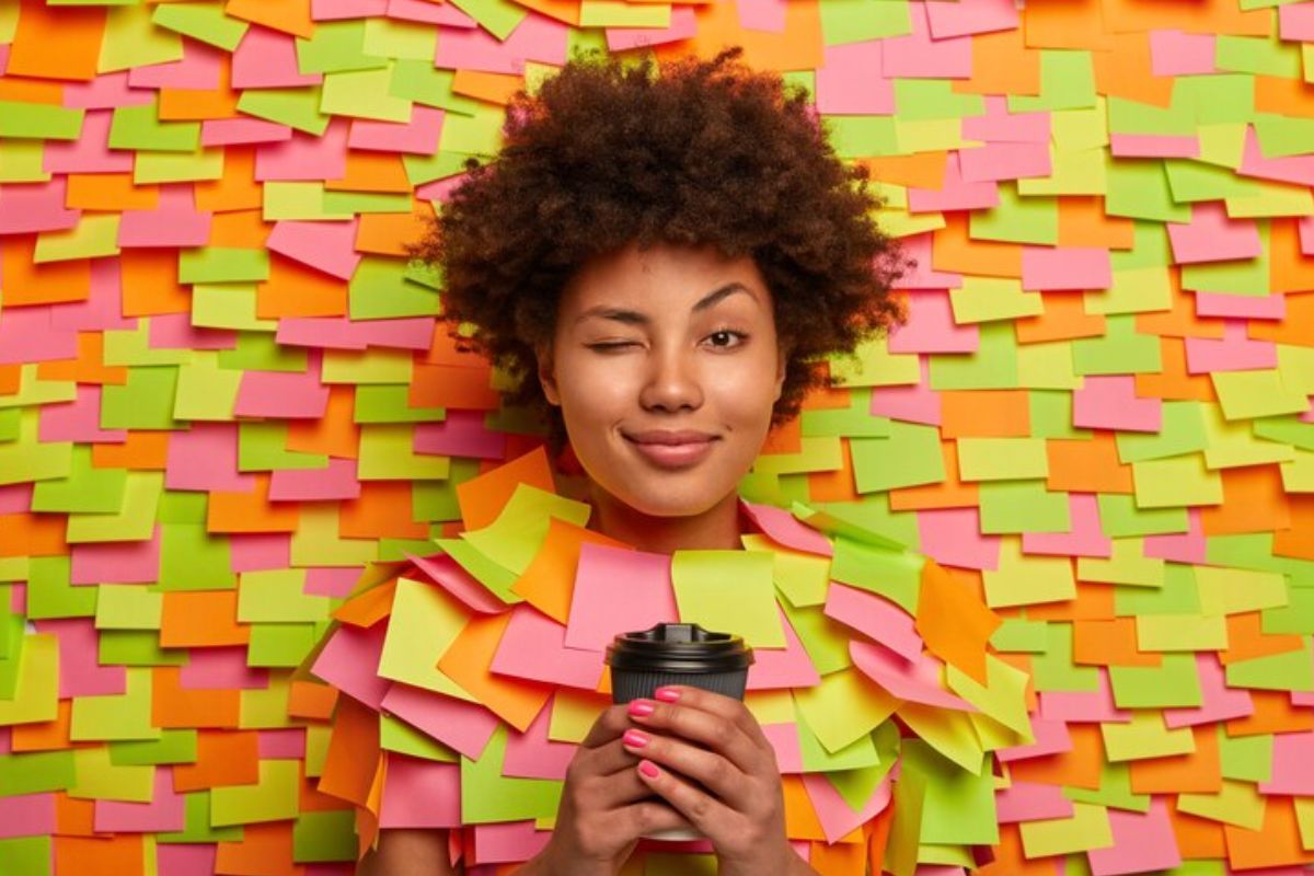 Como usar o Reminders para gerenciar tarefas diárias? Mulher jovem com cabelo afro sorrindo e piscando, segurando um copo de café, cercada por papéis coloridos de lembrete em tons de verde, rosa e laranja.
