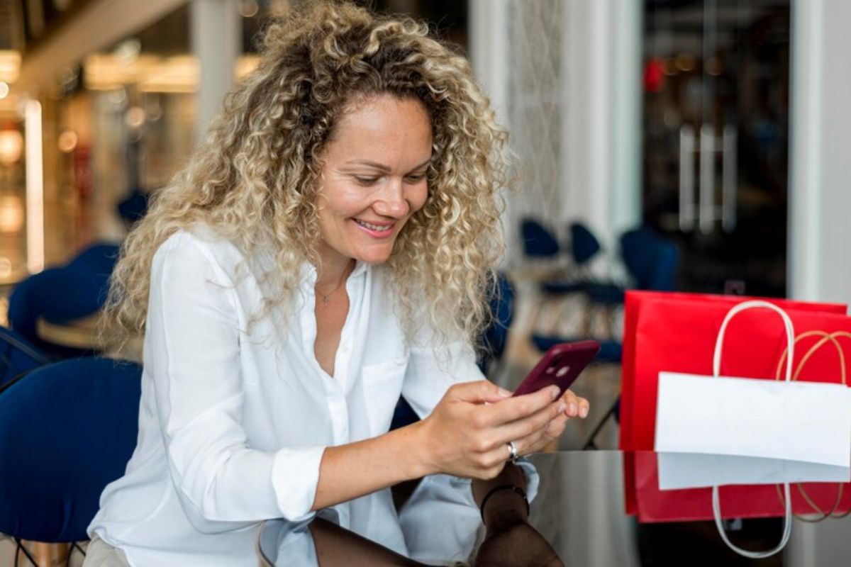 Dicas de mensagem para cliente para fechar mais vendas Mulher sorridente usando smartphone com sacolas de compras ao lado, sentada em um ambiente de shopping.