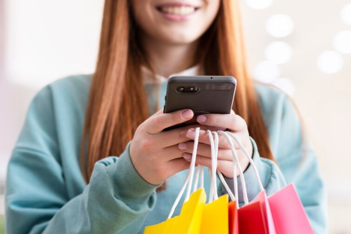 Tray: conheça essa plataforma virtual de vendas Mulher sorrindo enquanto segura sacolas de compras coloridas e usa um smartphone para verificar informações ou fazer compras online.