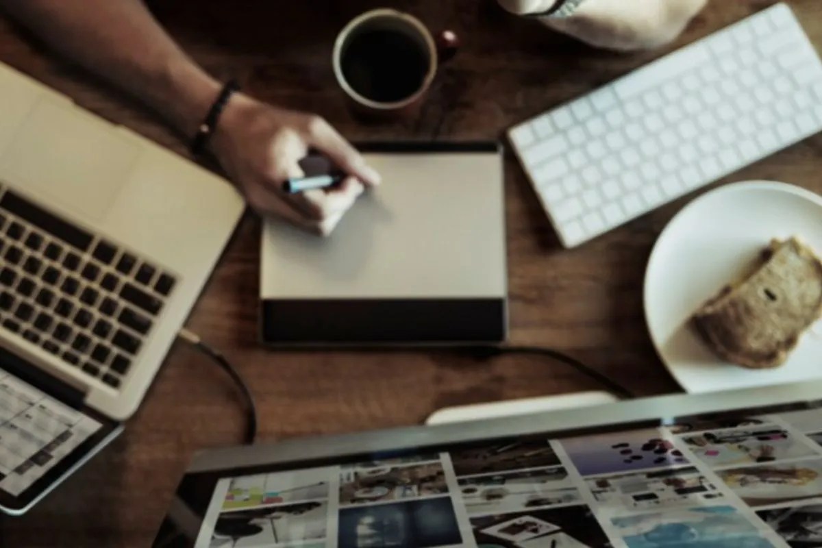 Como utilizar GIFs e animações para melhorar seus posts de blog Pessoa escrevendo em aparelho digital com pires à sua frente contendo um pedaço de pão e uma caneca de café ao lado, cercado por notebook e teclados sem fio, além de um monitor com diversos GIFs sendo exibidos na frente do indivíduo.