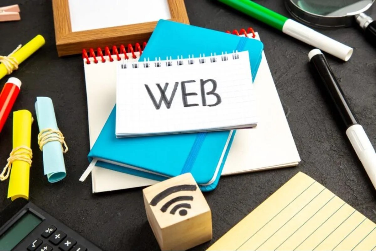 Entenda o que são sites PWA e como eles podem beneficiar seu negócio Mesa preta com objetos sobre a mesma em foto tirada de ângulo diagonal, mostrando alguns papéis enrolados e amarrados com barbante, um bloco de madeira com sinal de wi-fi desenhado neste em preto, com canetas hidracor de cores diversas, folhas e agendas espalhadas ao lado de uma calculadora e um caderno pequeno aberto com o nome WEB escrito, que faz analogia de elementos usados na criação de um site PWA.