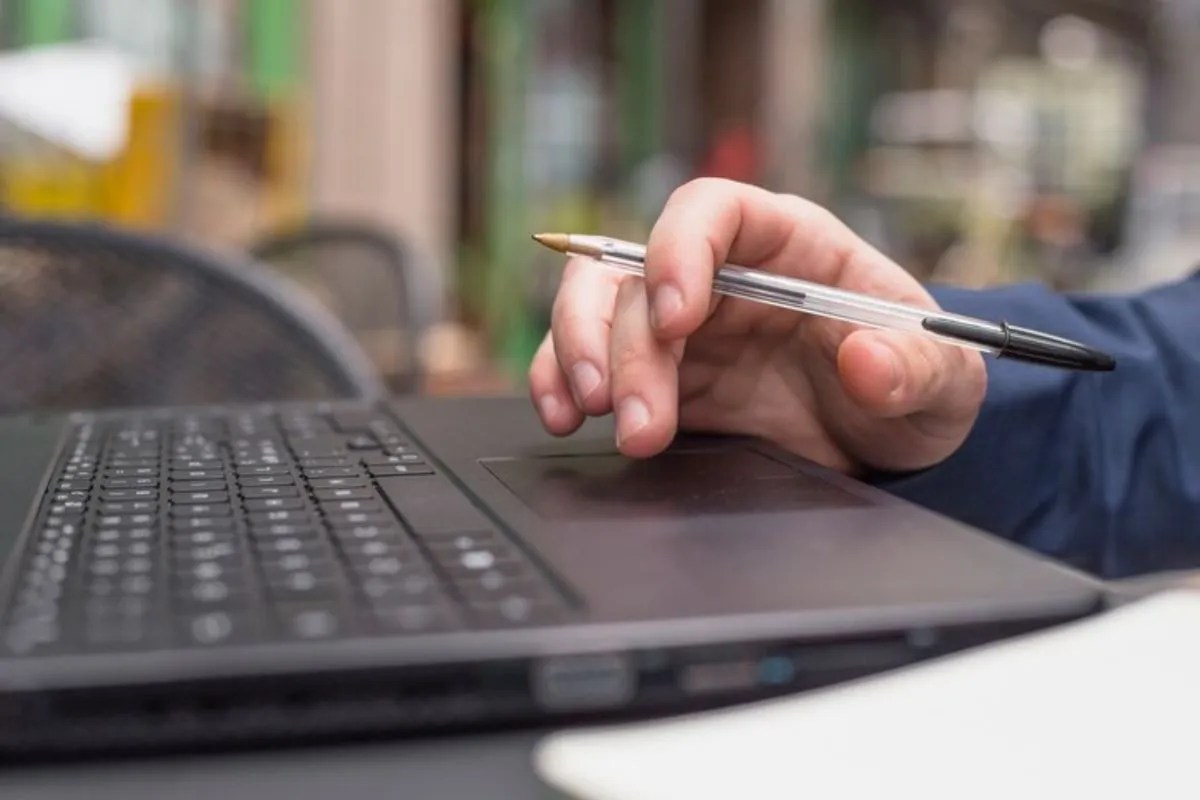 Mão de pessoa segurando caneta esferográfica simples e transparente com tampa e tinta pretas enquanto usa notebook.