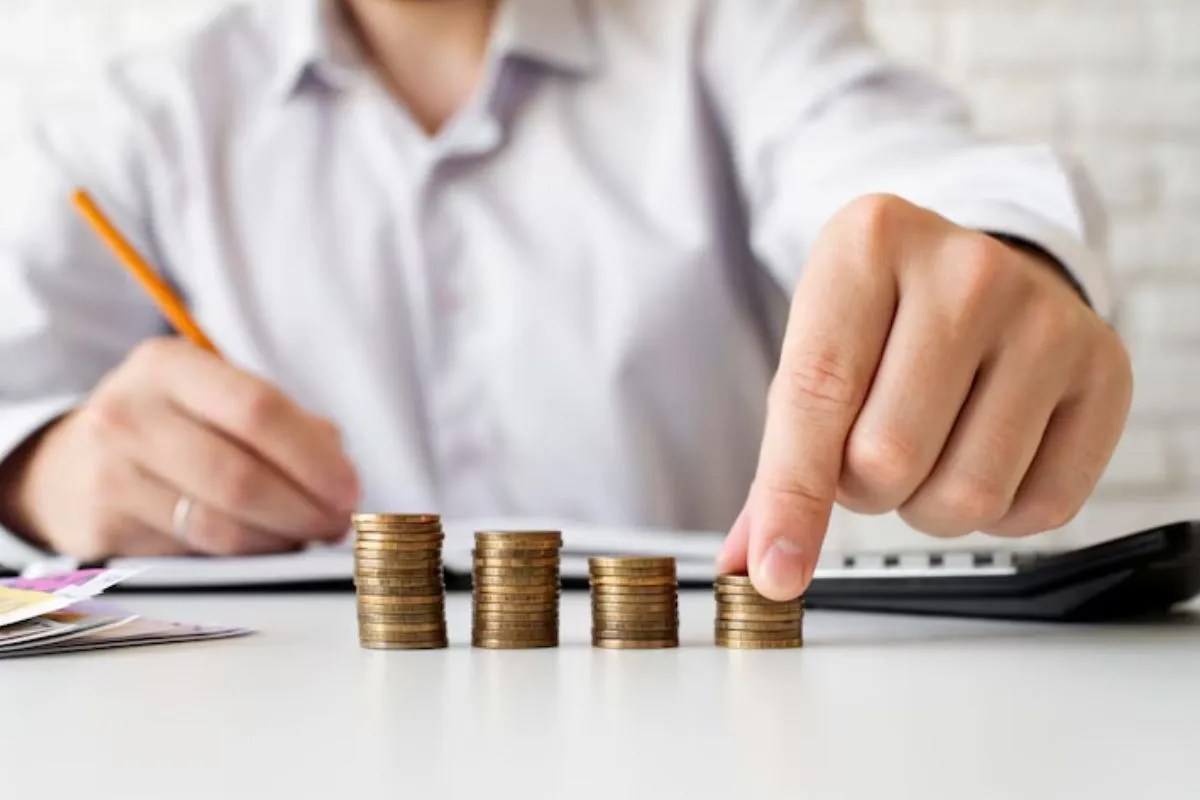 Negócios baseados em renda passiva: o que realmente funciona? Pessoa empilhando moedas sobre mesa branca enquanto faz anotações financeiras em papel ao fundo, em desfoque na fotografia.