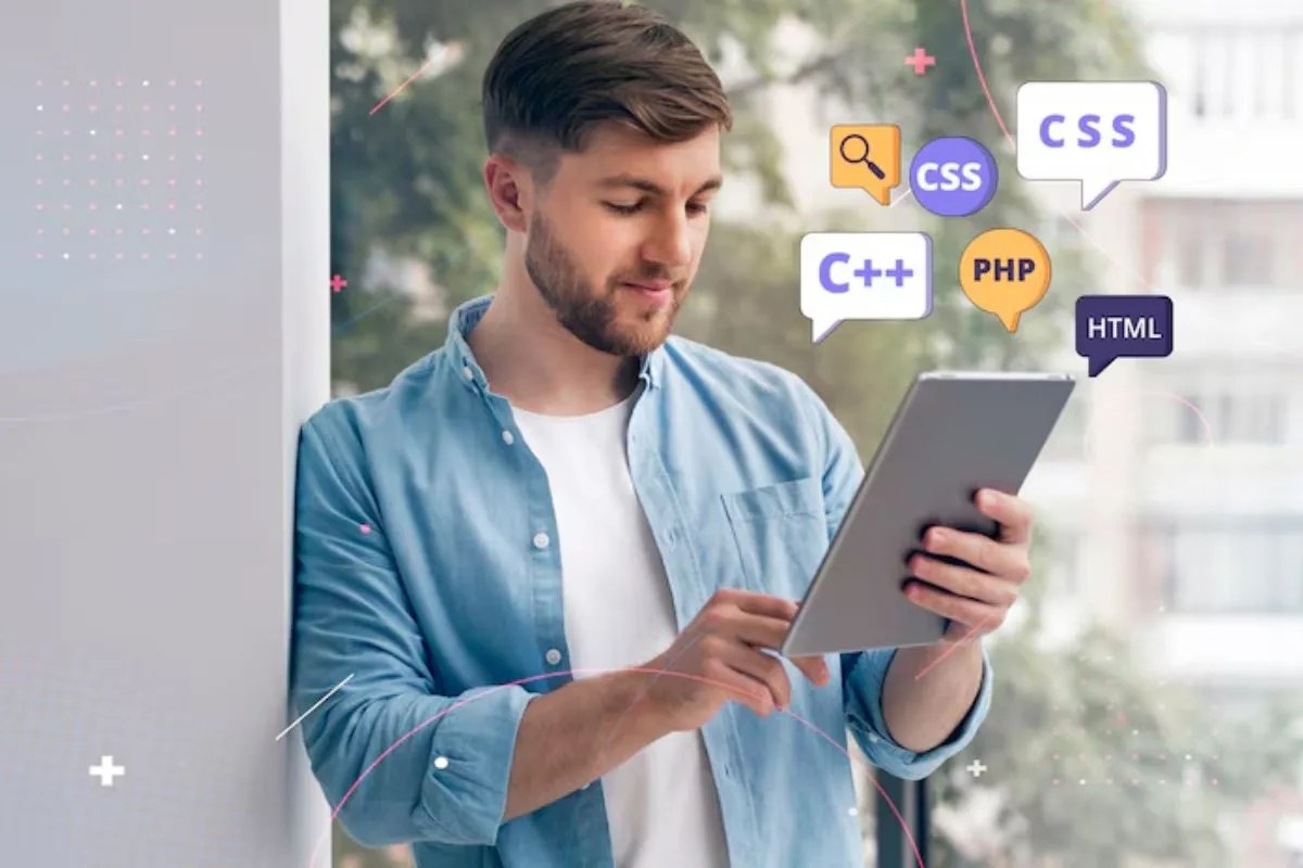 Homem de camiseta branca e camisa de botão azul aberta por cima encostado em parede branca usando um tablet na cor cinza, com alguns símbolos que fazem analogia à programação de sites PWA, além de algumas linguagens em montagem digital.