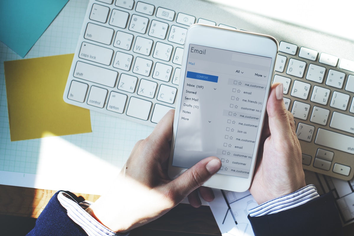 Pessoa segurando um smartphone com uma caixa de entrada de e-mails aberta, com um teclado de computador ao fundo.