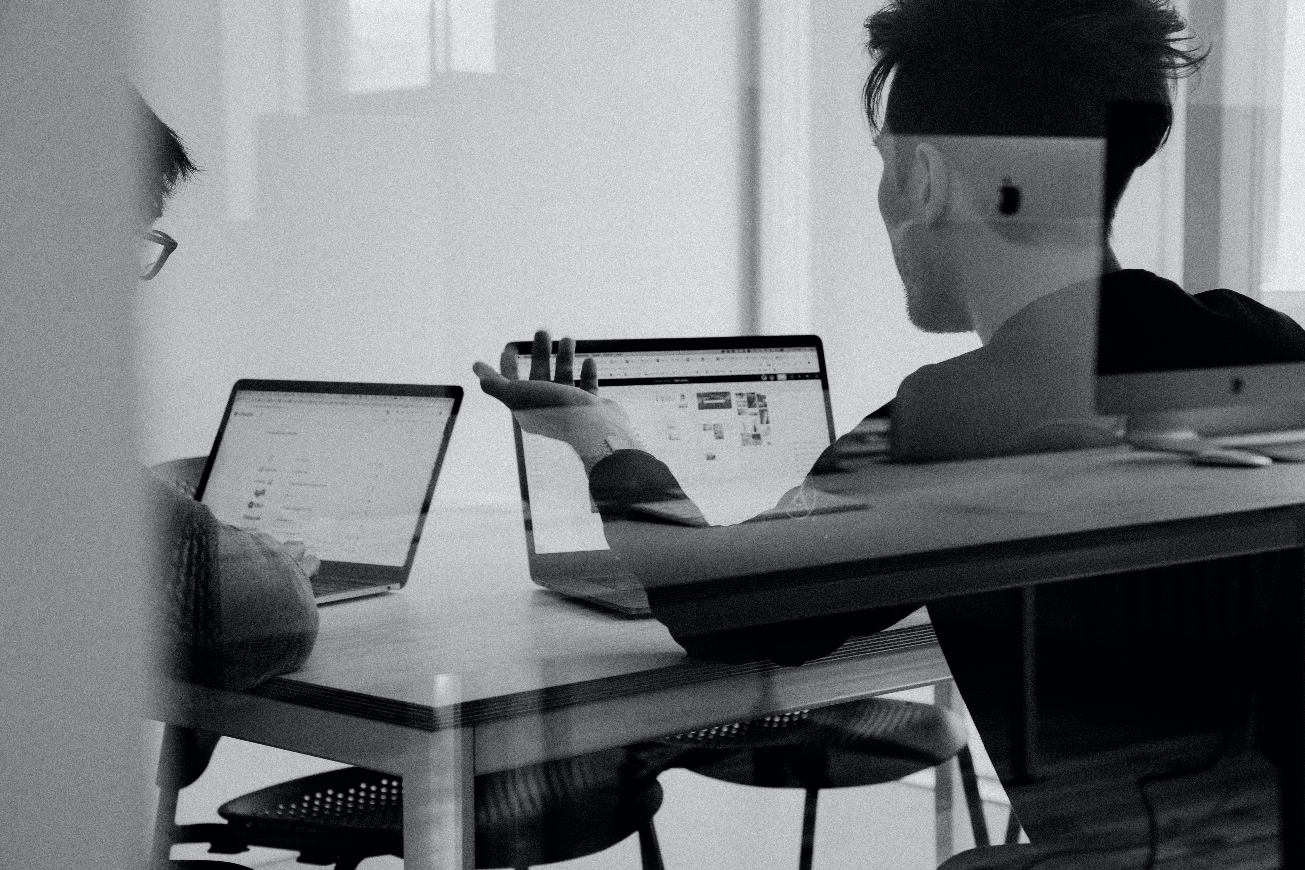 Two people with short hair look at computer screens with their backs to the camera; one is gesturing at something on the screen.