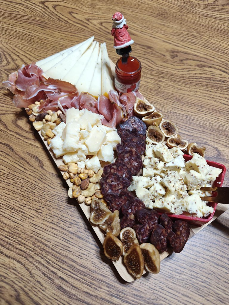 A medium sized board (you can't really see the board underneath everything, but it's a light wood board from Harry & David). There's triangular cut pieces of gouda, a river of prosciutto, a mound of crumbled cheddar, halved figs, scattered nuts, the red pepper jelly with a spreader utensil sticking out of the jar, the salami snaking its way around, and the Dragon Slayer crumbled in the corner, as well. 