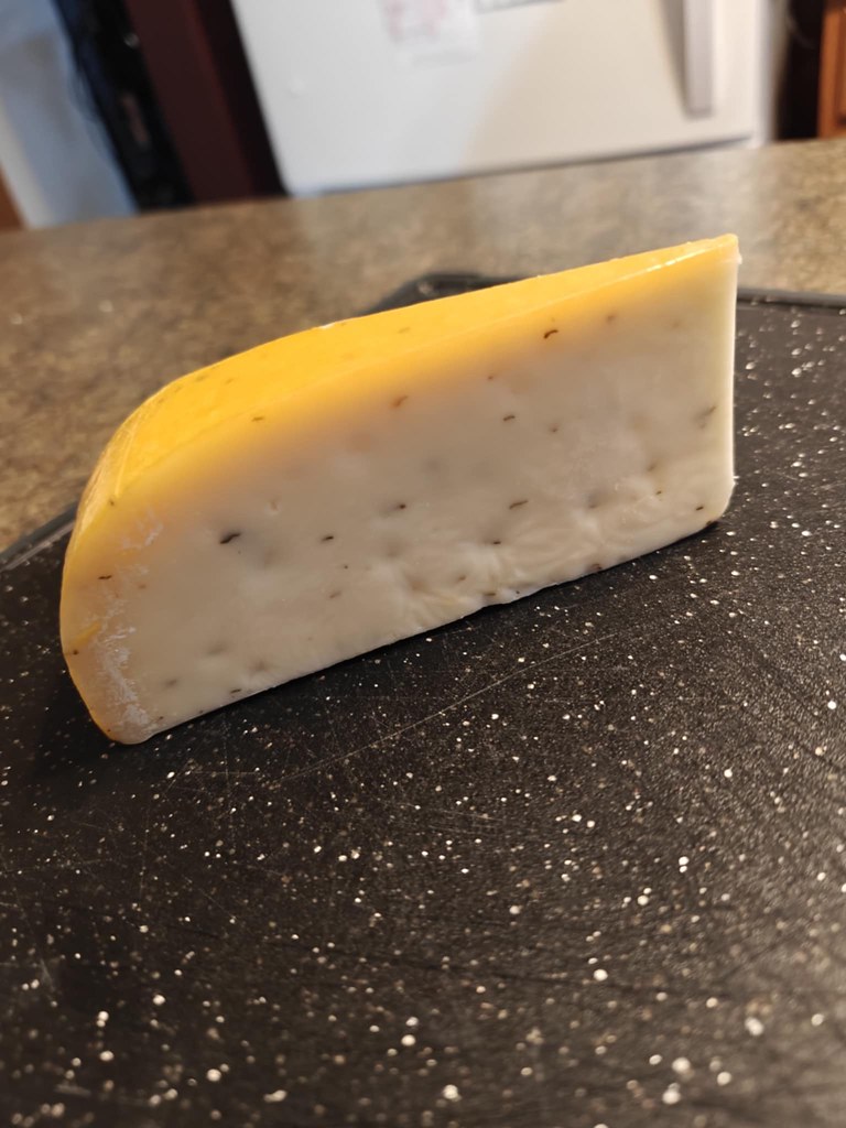 A small wedge of basil pesto gouda, an off-white in color with visible specks of herbs throughout the cheese. The rind is a pale yellow color. 