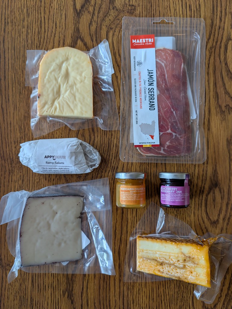 Everything from the box laid out on a wooden table. There's three cheeses, two meats, and two spreads. 