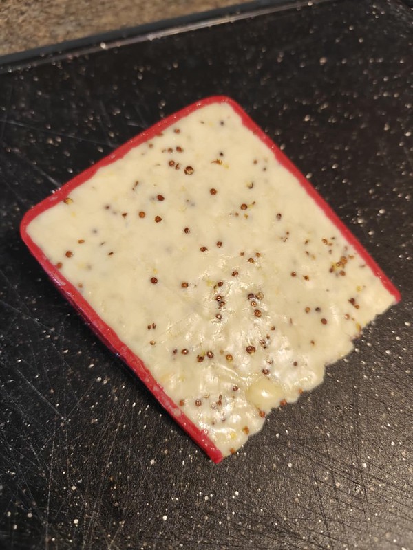 A wedge of Dragon Slayer cheese, an off white cheese with a ton of visible mustard seeds throughout it. The rind is a bright red wax, encasing the sides and top part. 