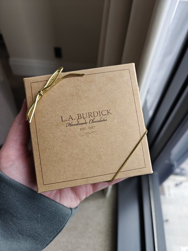 A little brown square box containing chocolates, the outside has the L.A. Burdick logo and a gold ribbon wrapped around the edges for decoration.