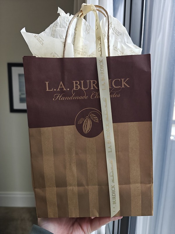 A brown bag from L.A. Burdick with their logo on it in a darker color, tissue paper sticking out of the bag, and a white ribbon tied around the handles for decoration.