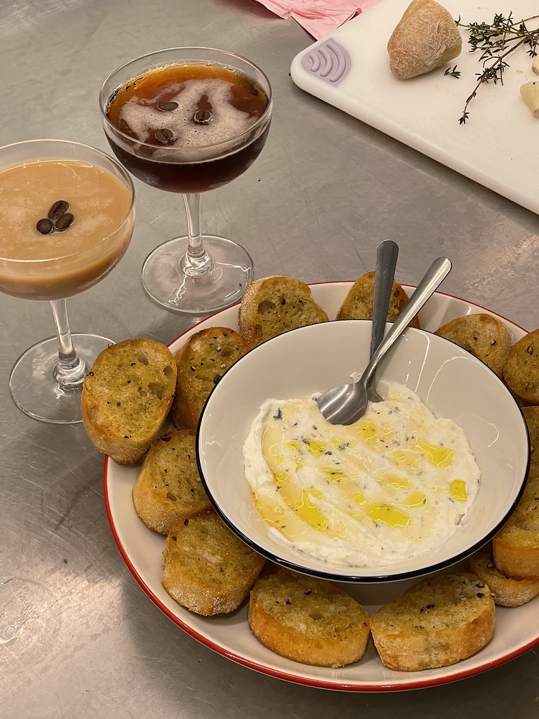 A small bowl full of olive oil drizzled ricotta, with crostini layered all around it. There's two espresso martinis visible in the shot, as well. One of them is much lighter brown in color than the other. Everything sits atop a stainless steel counter. 