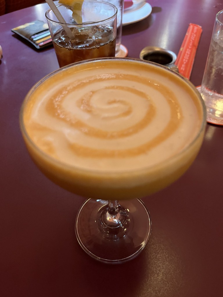 A coupe glass filled with an orange colored creamy liquid and a big white swirl of cream on top.