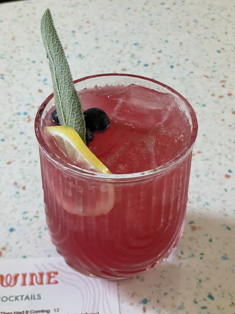 A short, textured glass filled with dark pink liquid and ice. The garnishes include blueberries, a lemon wheel, and a sage leaf. 