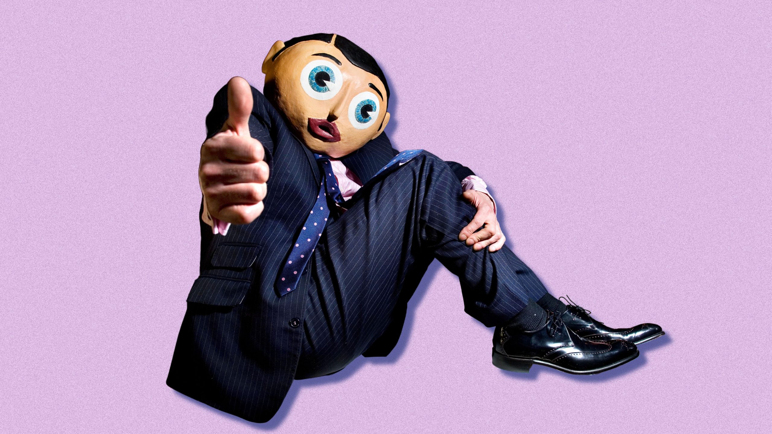 A man sitting in profile against a lilac-colored background, wearing a well-tailored pinstripe suit. He also has a giant plastic head in place of his actual head, and is giving a thumbs-up gesture to the camera.