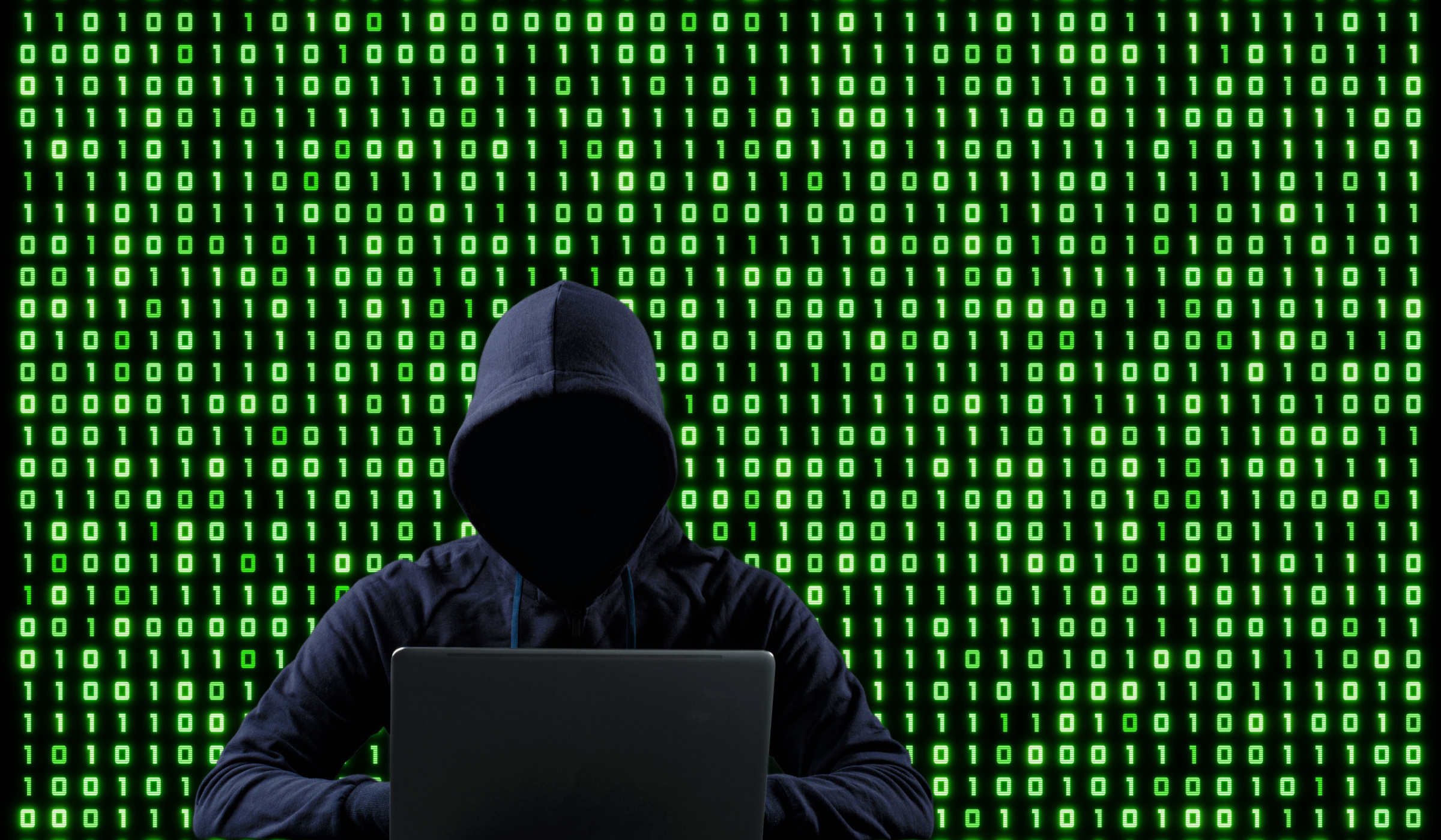 A person shrouded in hood sits at a computer, against a backdrop of green binary code