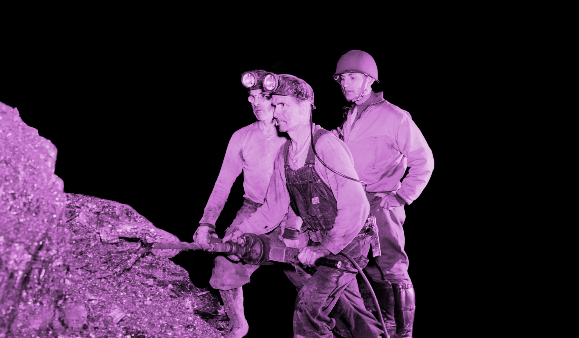 A miner drills into a rock formation while two men wearing headlamps look on