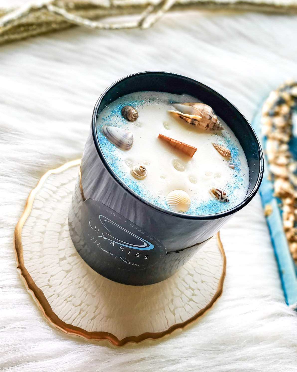 Moonlit Shores soy candle in a black glossy tumbler jar, featuring a wooden wick and decorated with natural seashells and soft-toned botanicals.