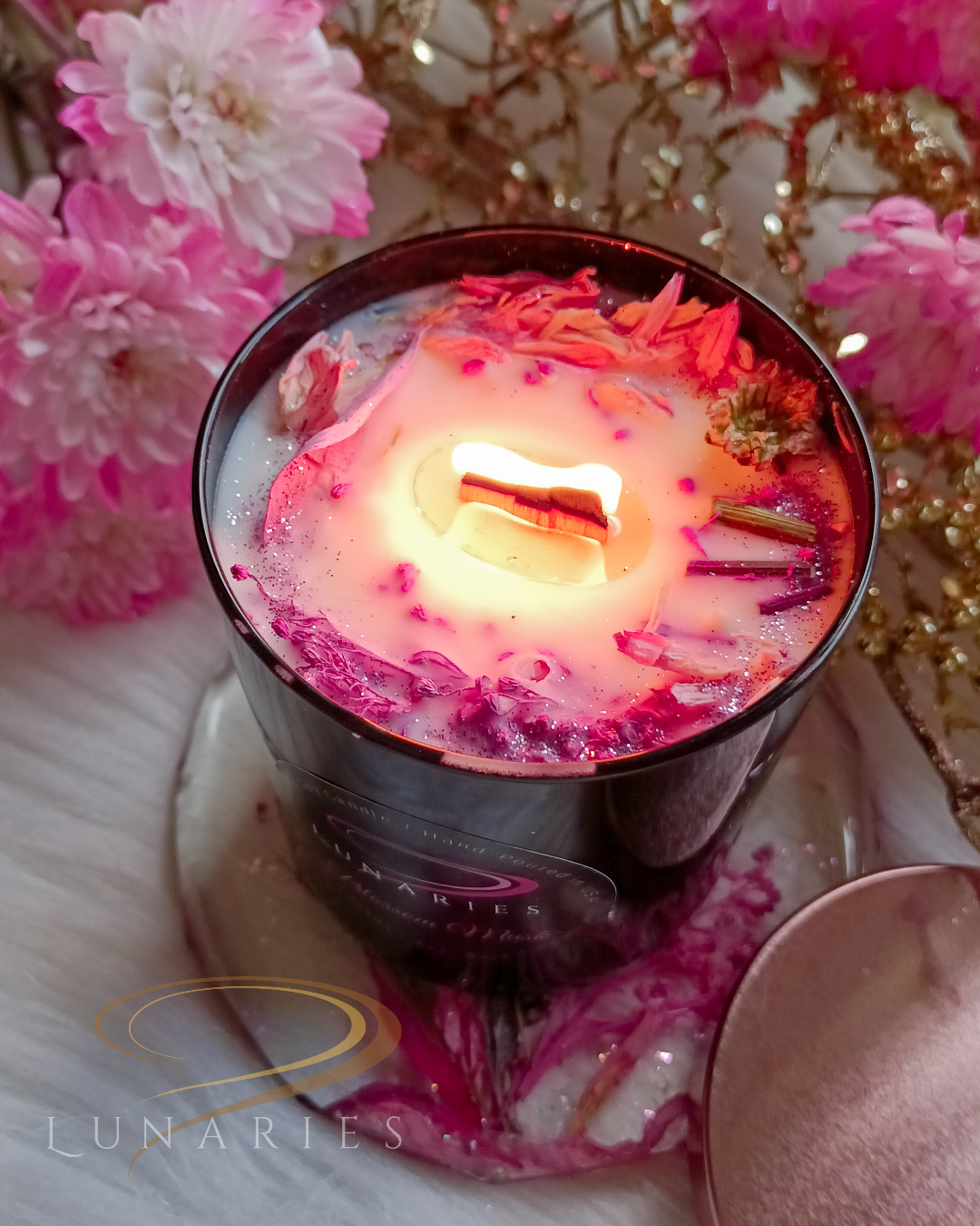 pink burning candle with wooden wick in a sleek black tumbler jar with pink botanicals on the top