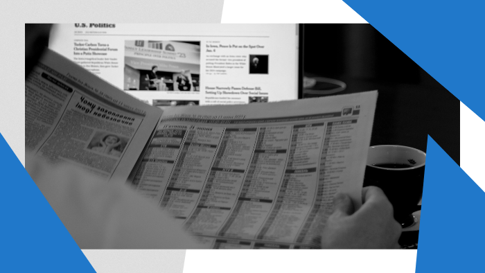 Person reading a newspaper in front of a laptop with news on the screen