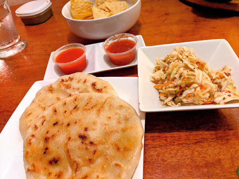 A plate with naan bread, two bowls containing salsa and chips, and a side of coleslaw on a wooden table.
