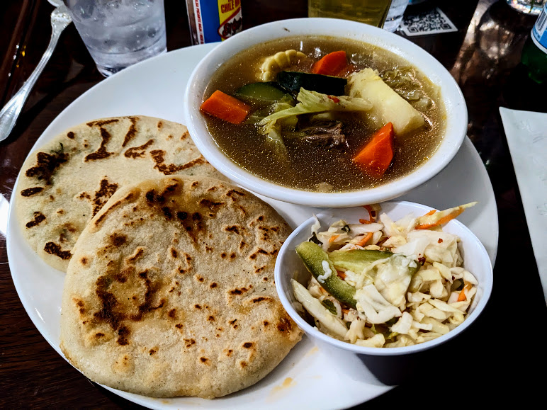 A meal consisting of two pupusas, a bowl of soup with vegetables and beef, and a side of coleslaw on a wooden table.