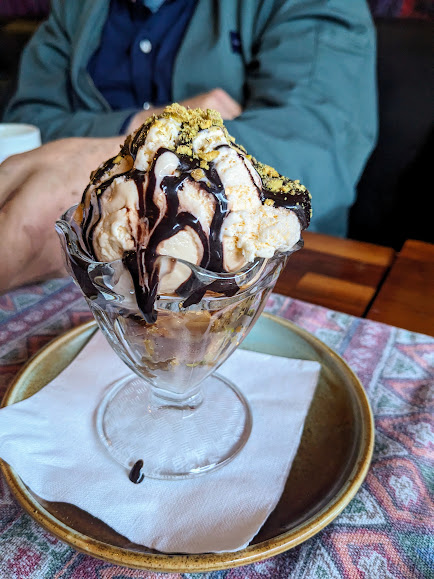 A person sitting behind a glass bowl of ice cream sundae with chocolate syrup and sprinkles.