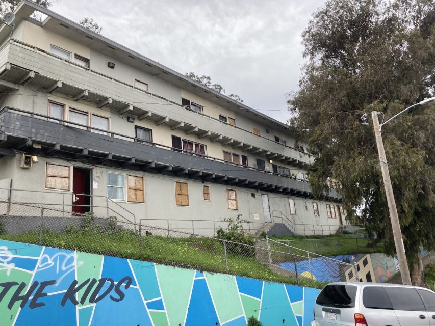 A multi-story building with boarded-up windows behind a chain-link fence with graffiti on a wall that reads "the kids.