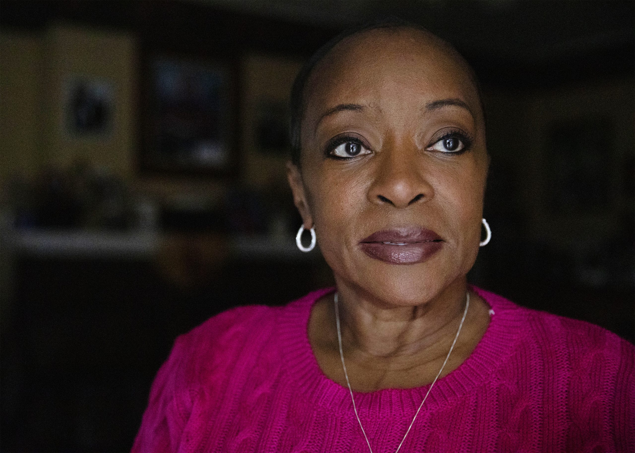 A Black woman stands for a portrait. She is wearing a silver hoop earrings, a silver chain, and a knit fushia sweater.