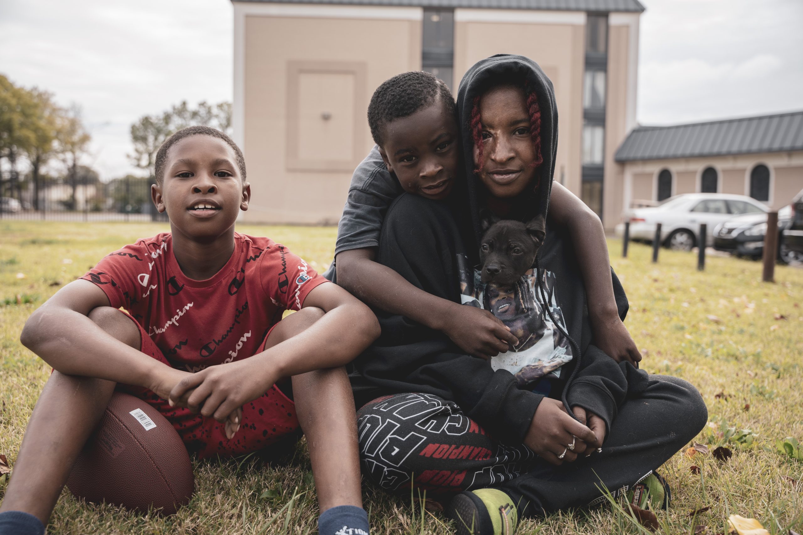 A Black woman wearing a hoodie with a small dog peaking out of the neck sits in the grass with two small Black boys. One boy is seated to her left. The other boy is draped across her back.