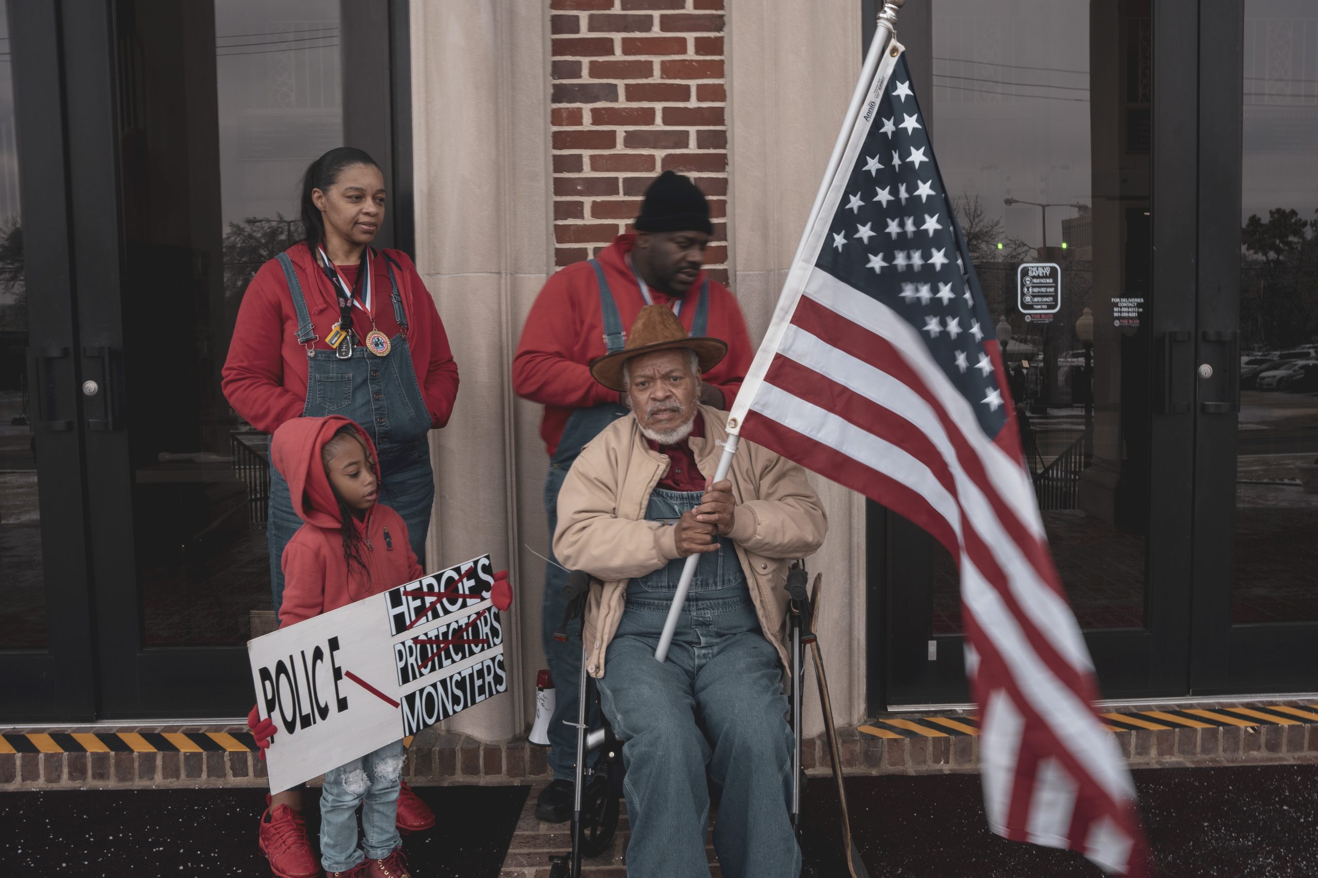 Four Black people wait outside a building. One is seated holding a large American flag.