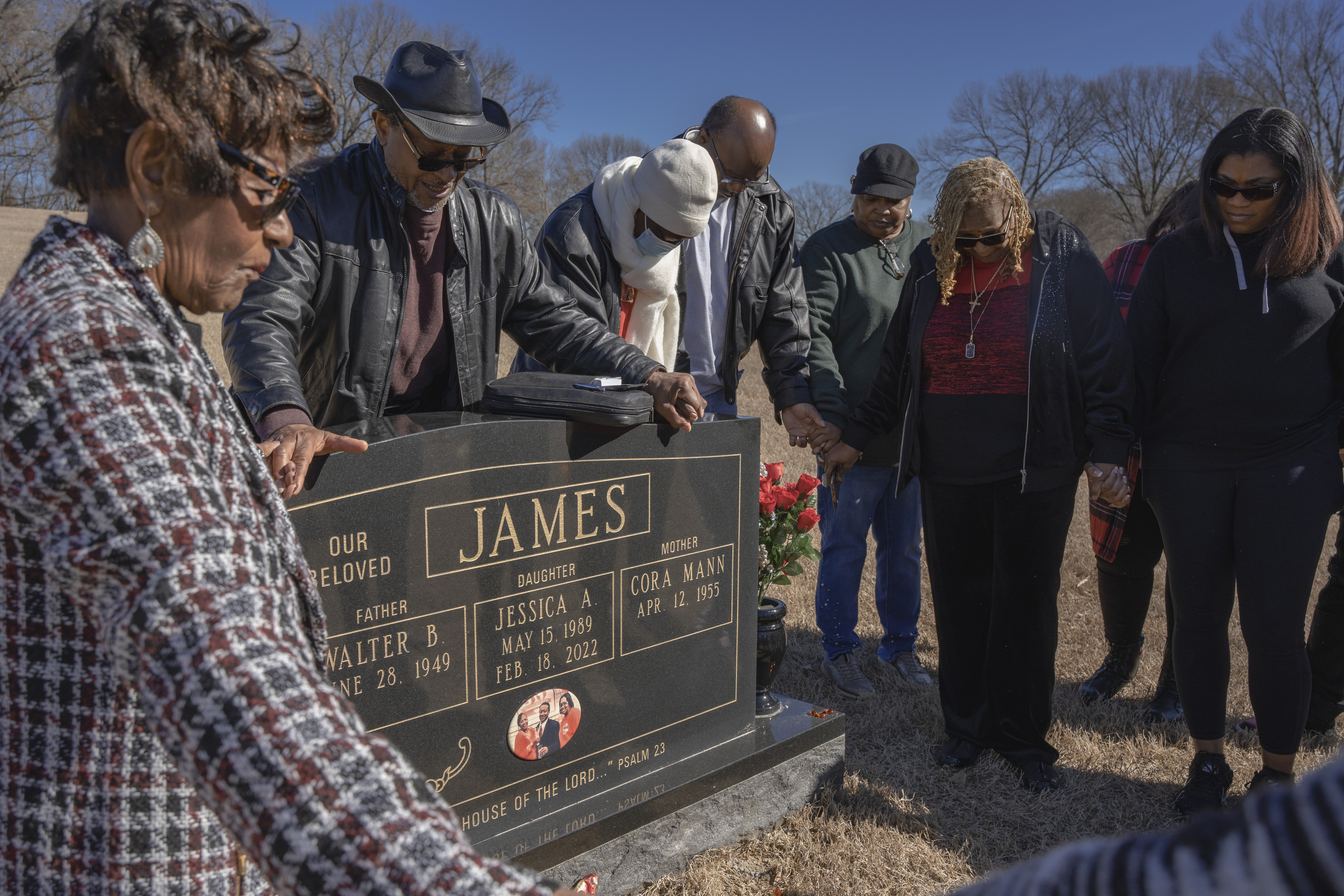 A group of people hold hands and bow their heads around a headstone. 