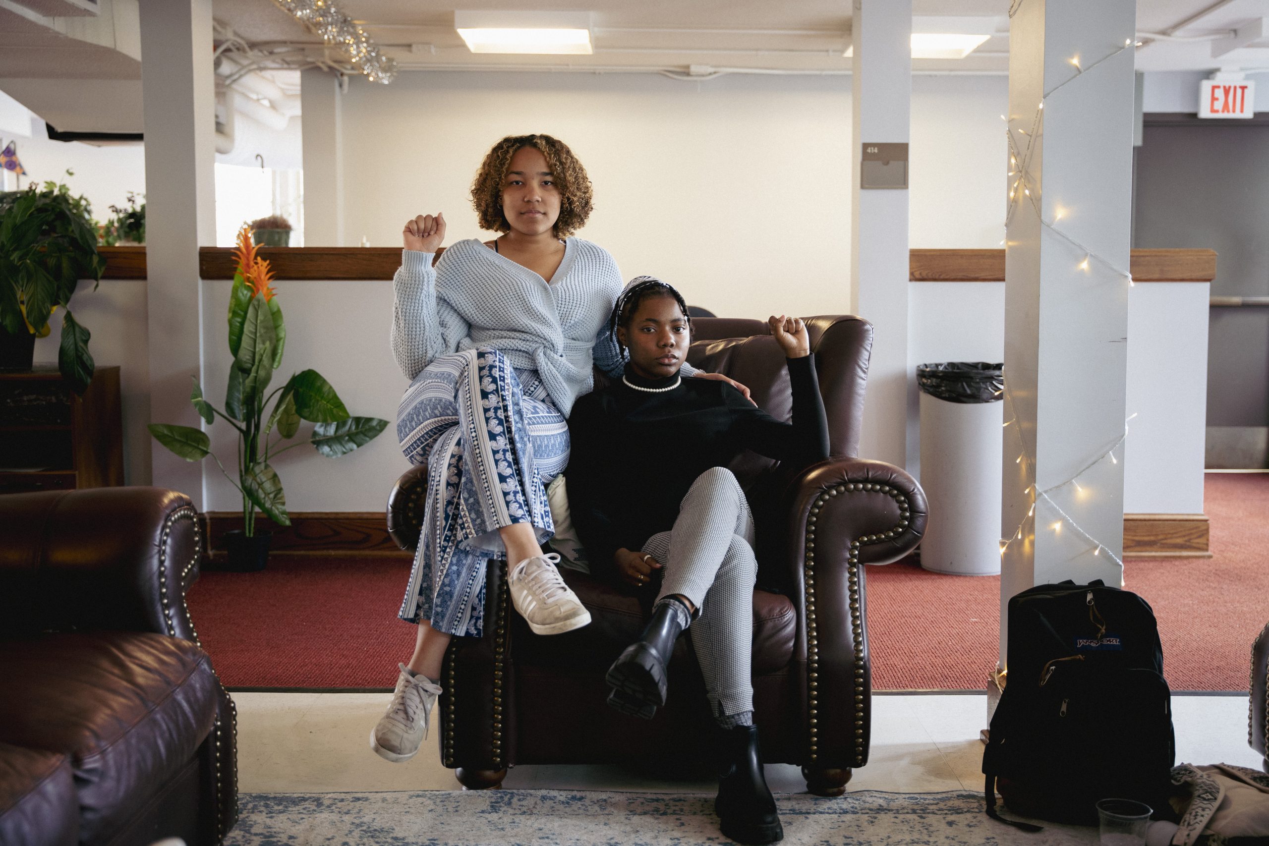 Two Black hold their fists in the air while seated on a brown leather chair inside Rhodes College's Interfaith Lounge.