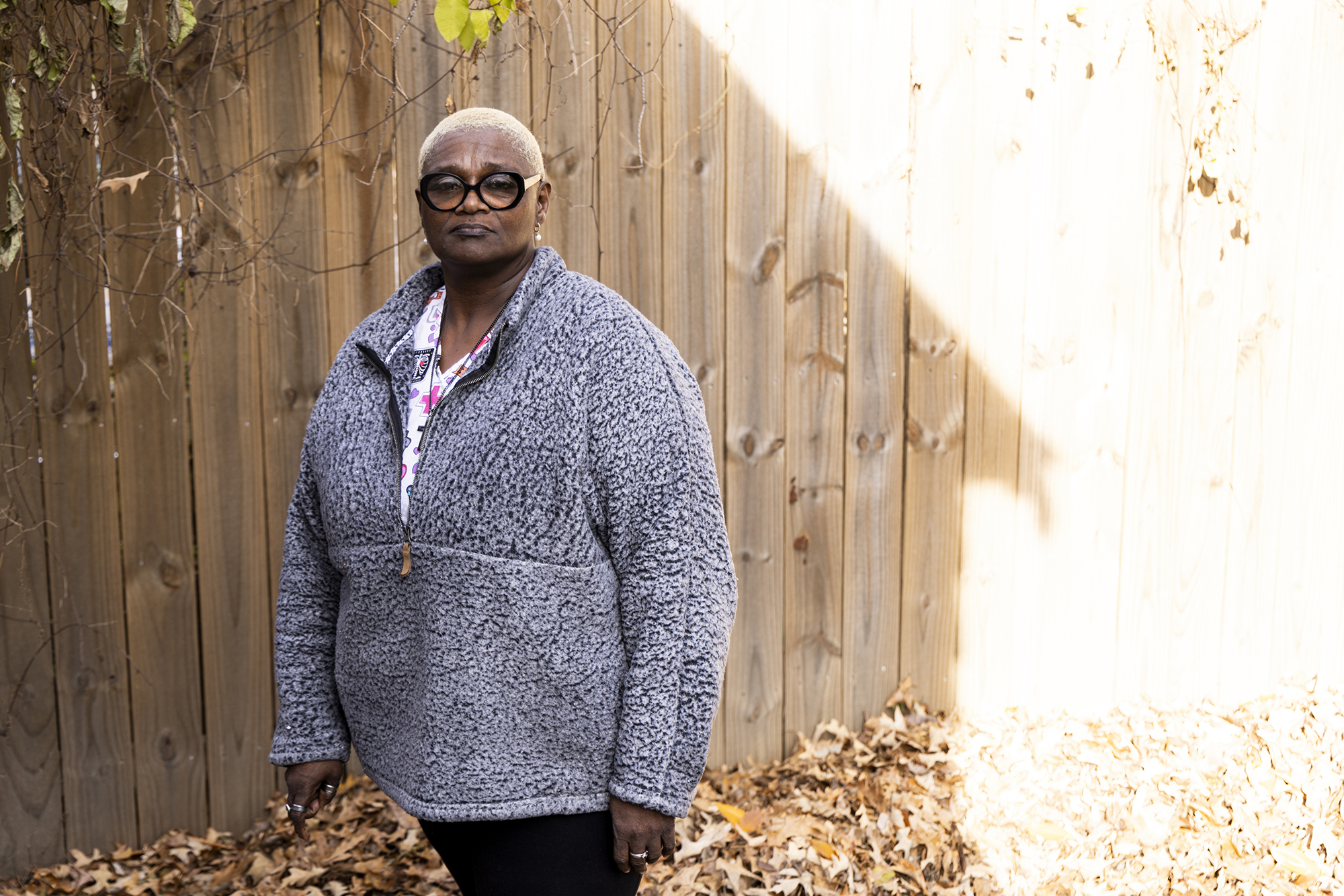 A Black woman stands in front of a wooden fence with piles of fall leaves at her feet. 