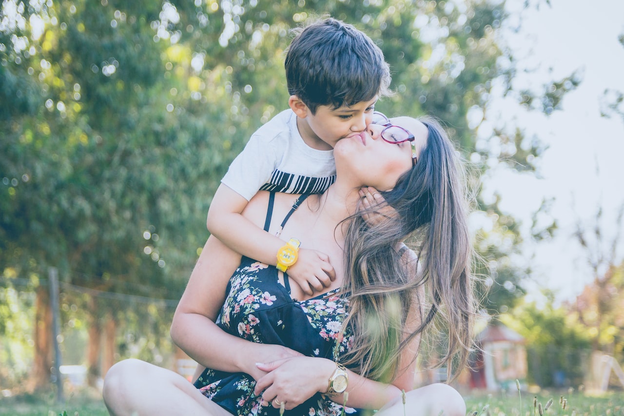 Woman sitting on ground while boy standing on her back kissing her at daytime