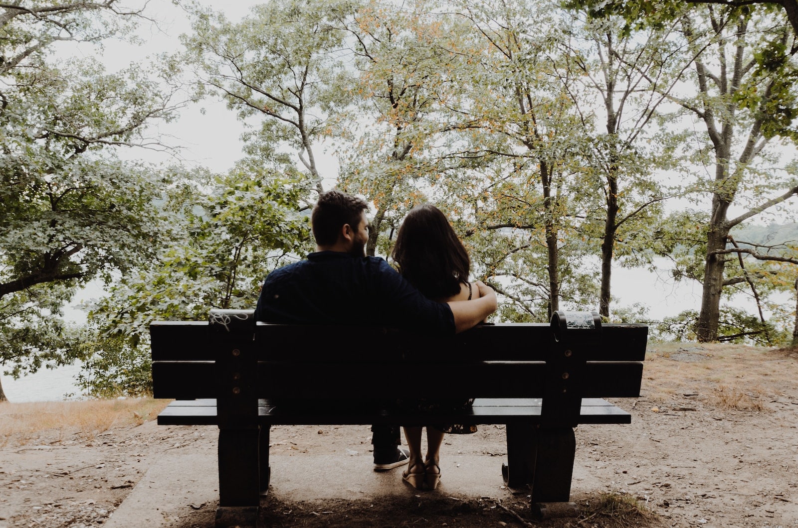 Man looking to woman sitting on black wooden bench in front of tall trees during daytime