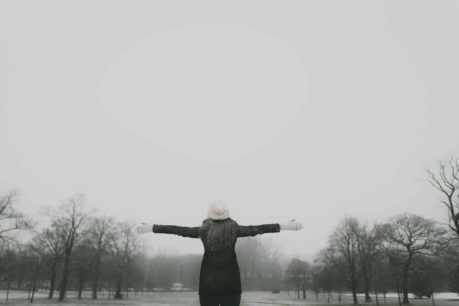Woman in black long-sleeved shirt standing and facing trees with stretched arms under white sky-life is crazy