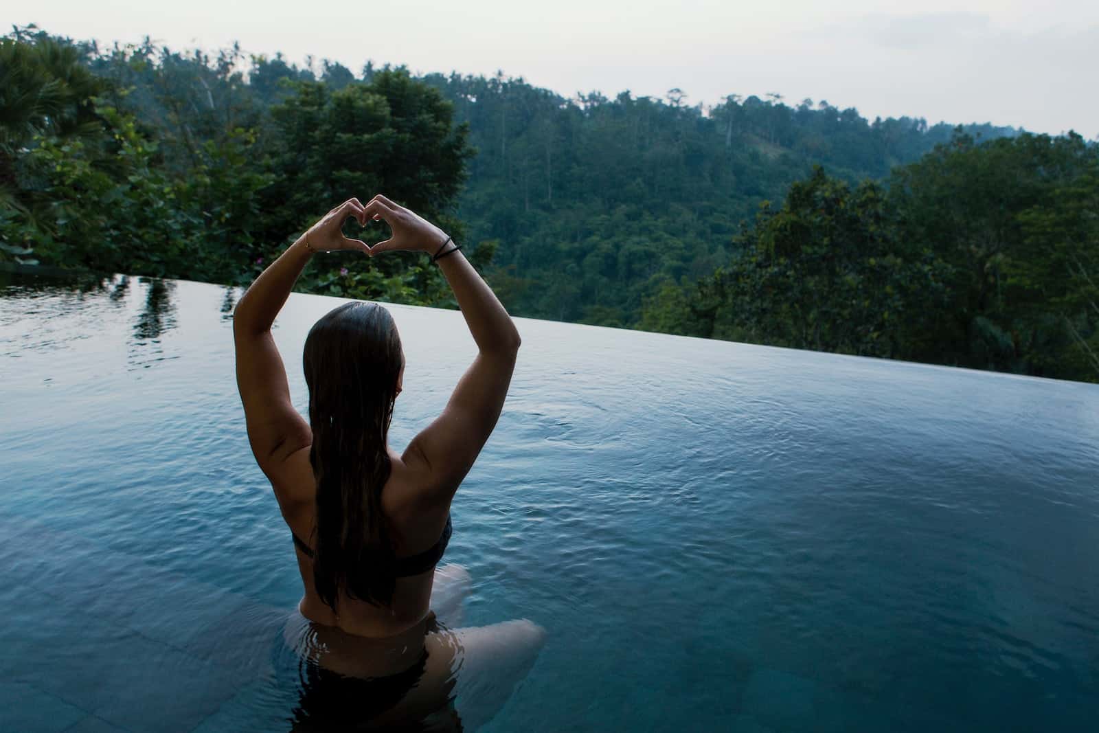 Woman in infinity pool making heart hand gesture facing green leafed trees-holistic health