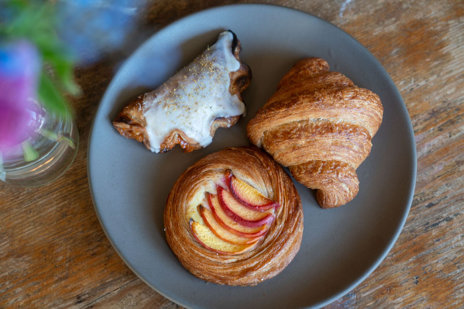 Three pastries, an apricot pop tart, peaches and cream danish and croissant sit on a gray plate next to a vase of flowers.