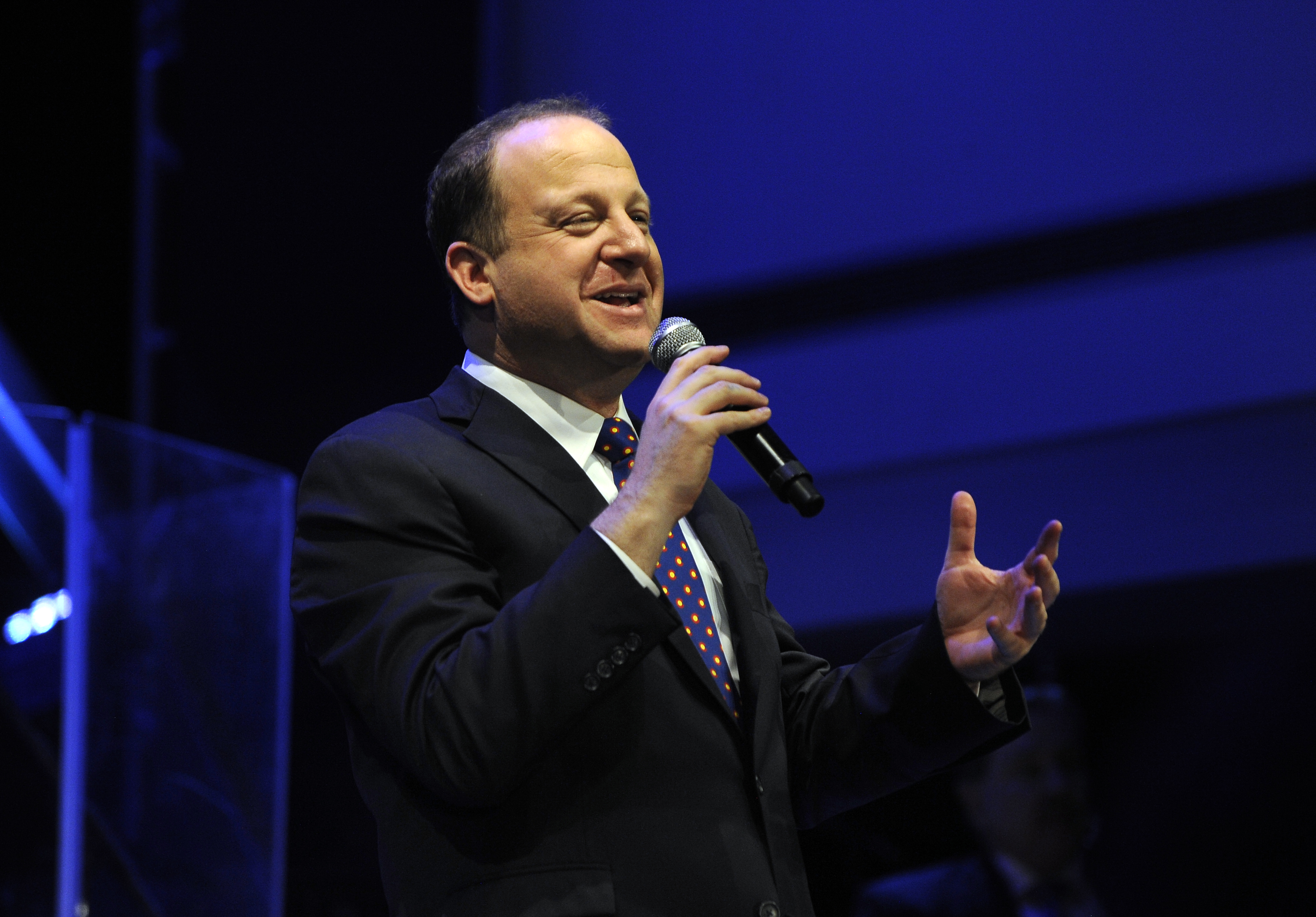 Gov. Jared Polis, wearing a suit and tie, speaks into a microphone on a stage with blue lighting.