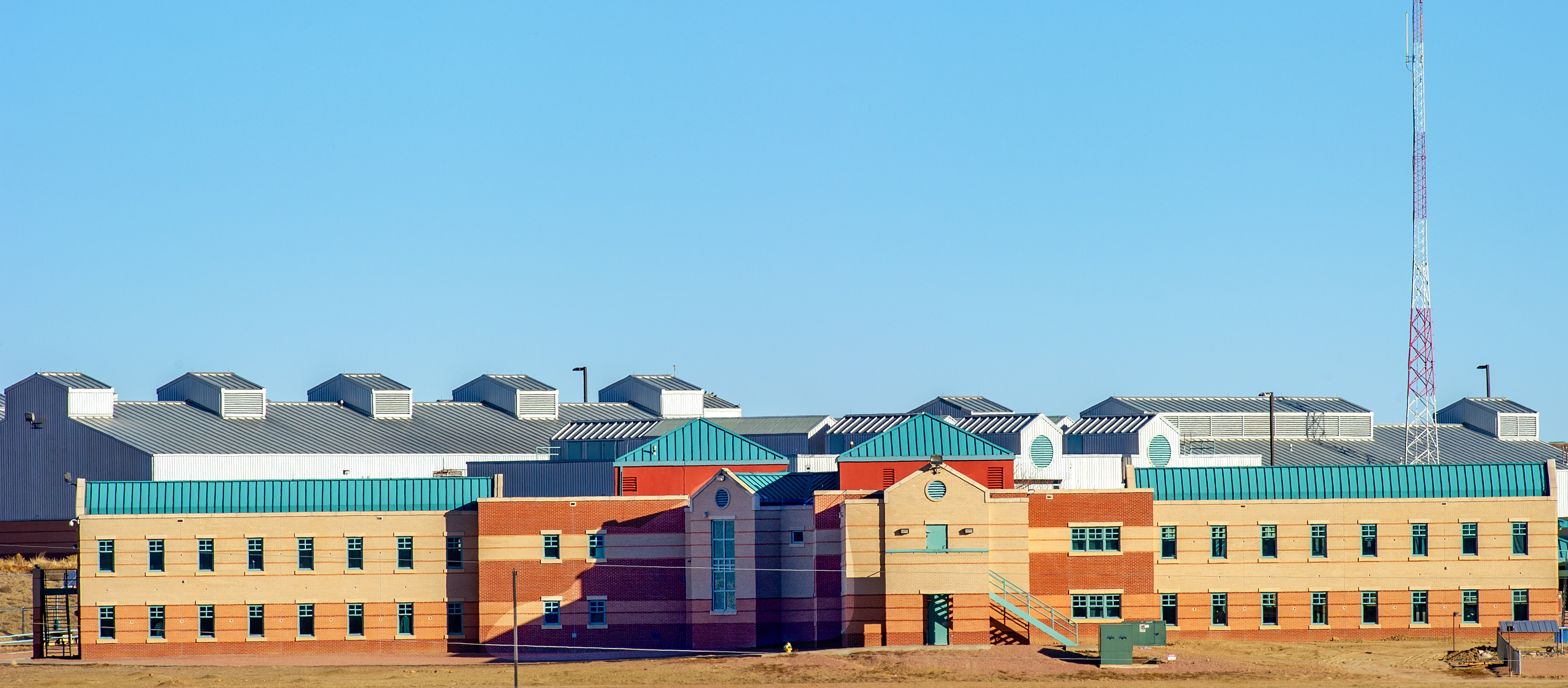 The Federal Correctional Complex, a large, modern brick and metal building with sections of blue roofs stands under a clear blue sky, featuring many windows and a tall communication tower to the right.