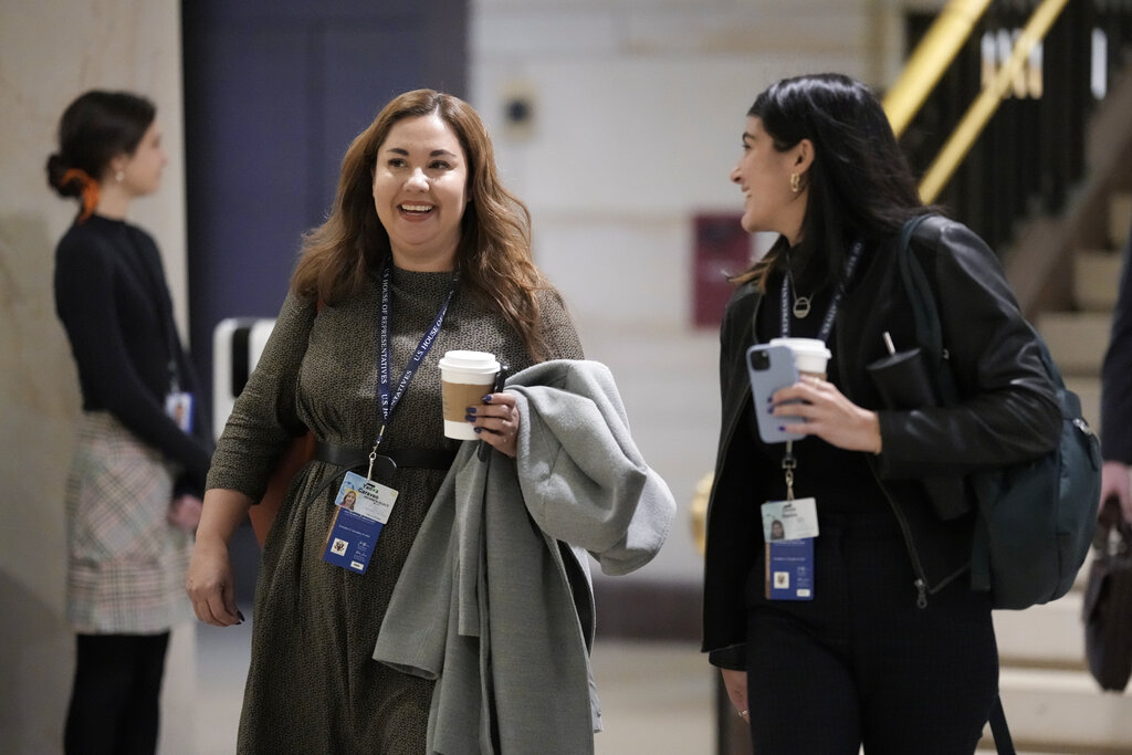 Two women walking indoors, each holding a cup of coffee and wearing ID badges. One woman is carrying a coat in her arm.