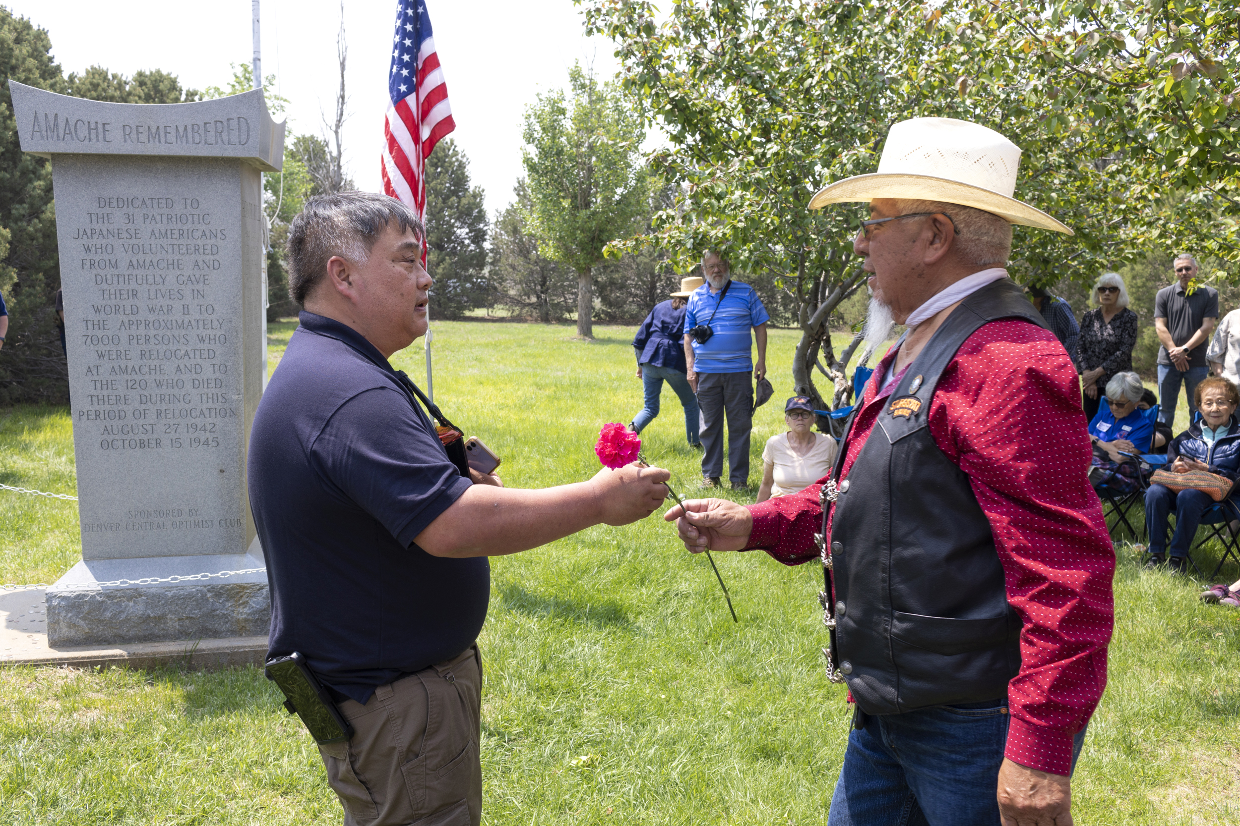 A man hands a flower to another man in front of a stone monument.