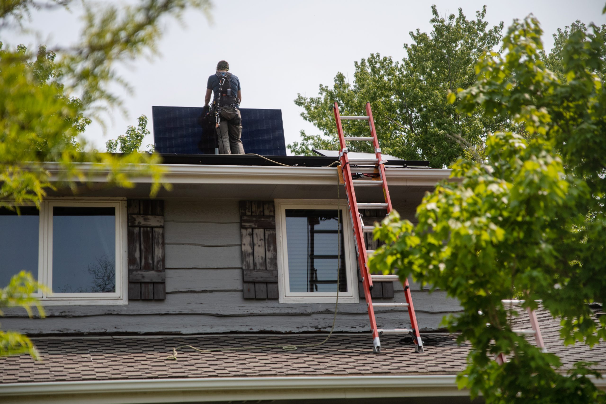 A worker installs solar panels on the roof of a residential home, accompanied by a red ladder extending from the ground to the roof.