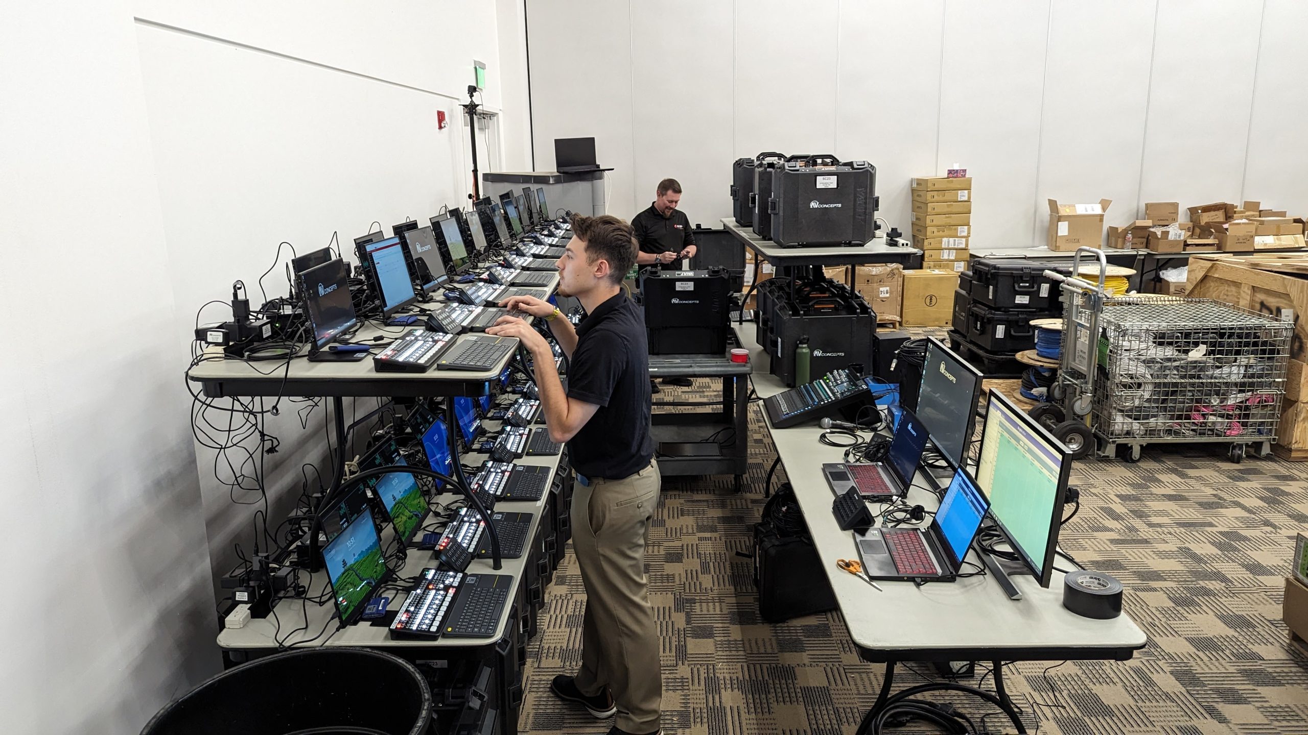 A table and shelf above it are covered in computers lined next to each other. A man stands while typing on one of the computers on the shelf.