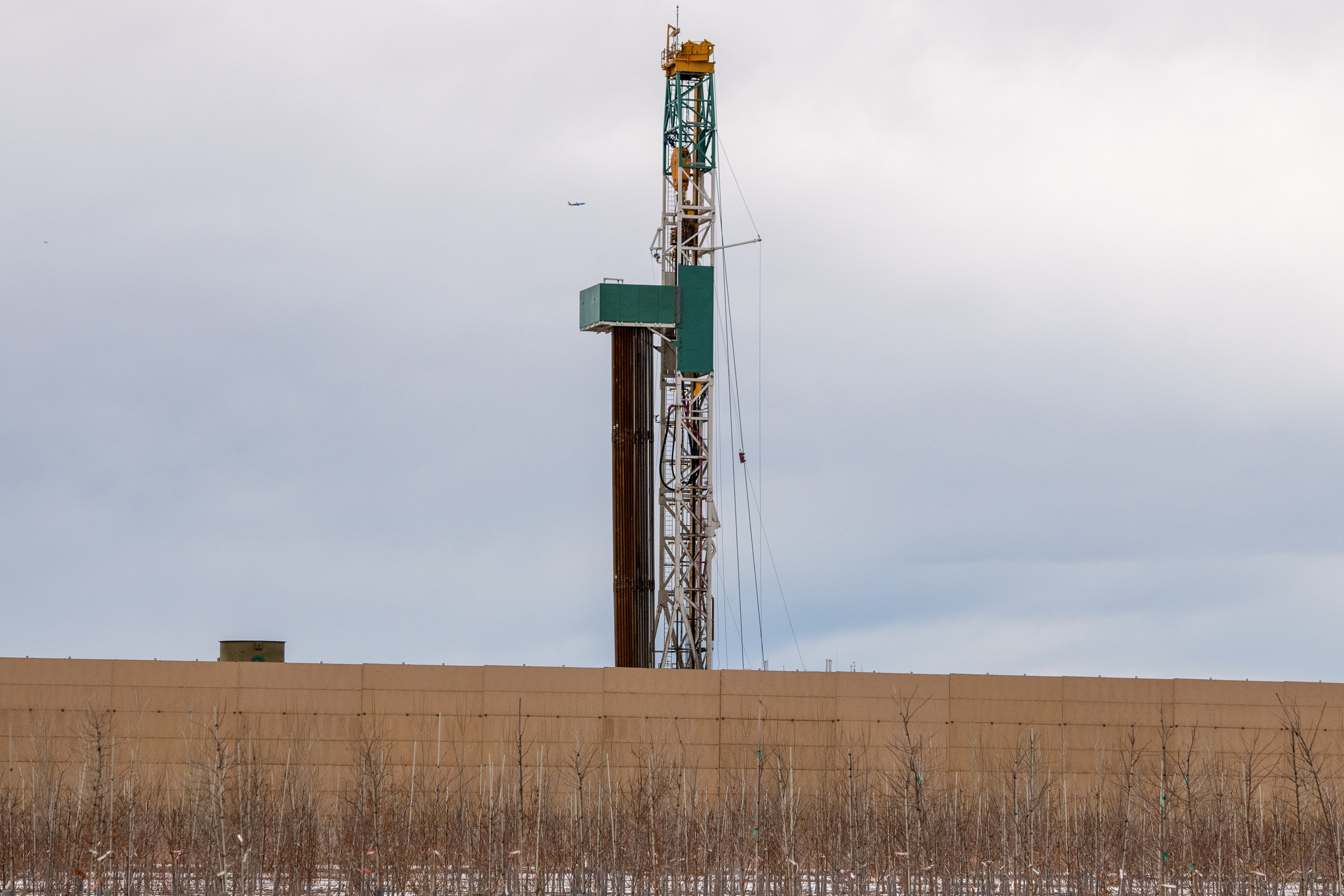 An oil drilling rig behind a concrete wall with sparse vegetation in the foreground under a cloudy sky.